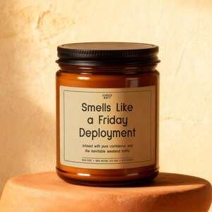 May include: A brown glass jar candle with a black lid sits on a terracotta-colored pedestal. The candle's label reads "Smells Like a Friday Deployment." Eucalyptus leaves are in the background. The candle is infused with pure confidence and the inevitable weekend vibes.