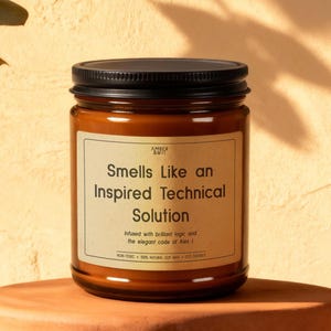 May include: A brown glass jar candle with a black lid. The label reads "Smells Like an Inspired Technical Solution." The candle sits on a small, terracotta-colored stand, with a light-colored background and leafy sprigs.