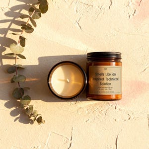 Top-down flat lay view of the funny programmer candle next to its black metal lid, styled with eucalyptus branches on a textured surface.