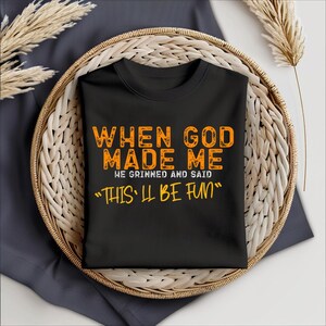 May include: Black t-shirt with orange text that reads "When God Made Me He Grinned And Said "This'll Be Fun""