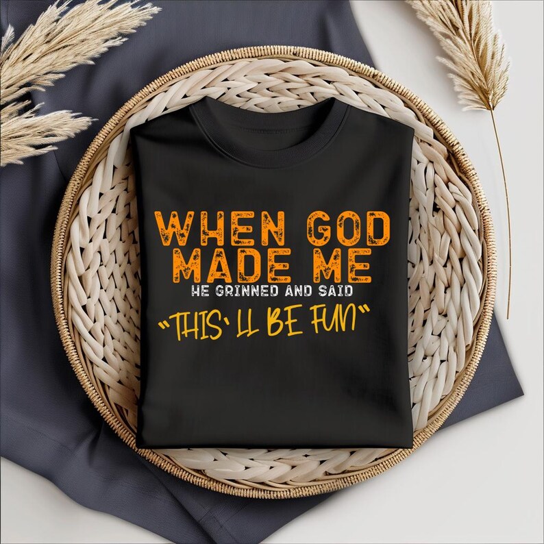 May include: Black t-shirt with orange text that reads "When God Made Me He Grinned And Said "This'll Be Fun""