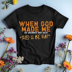 May include: Black t-shirt with orange text that reads "When God Made Me He Grinned And Said "This'll Be Fun""
