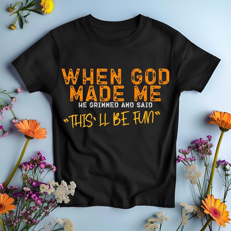 May include: Black t-shirt with orange text that reads "When God Made Me He Grinned And Said "This'll Be Fun""