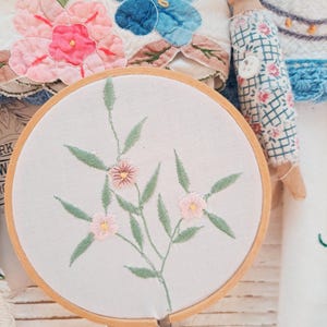 May include: A close-up of an embroidery hoop with a floral design. The embroidery features a light-colored fabric with a delicate pattern of pink flowers and green leaves. The wooden hoop holds the fabric taut, showcasing the detailed needlework.