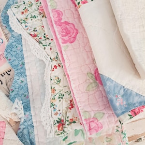 May include: A collection of layered fabric scraps featuring various patterns including pink roses, small floral prints, and blue geometric designs. The pieces are quilted with white stitching and accented with white lace trim and solid white fabric sections.