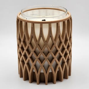 Puede incluir: Una vela en un tarro de cristal transparente, encerrada en un enrejado decorativo de madera marrón. El enrejado tiene un patrón de diamantes repetido. La vela es blanca y parece sin perfume. El portavelas cilíndrico se encuentra sobre una superficie blanca.