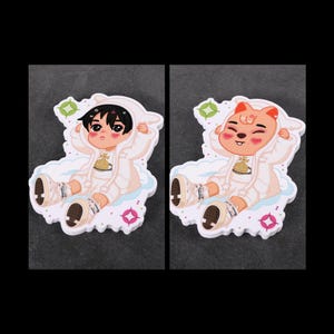May include: Two stickers featuring cartoon characters in white hooded outfits, sitting on clouds. One character has black hair, the other a bear-like face. Both are wearing sneakers and surrounded by decorative shapes.