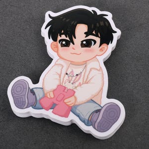 May include: A sticker featuring a cartoon illustration of a seated figure with black hair, wearing a white fuzzy jacket, light blue jeans, and purple shoes. The figure is holding a pink object. The sticker has a white border.