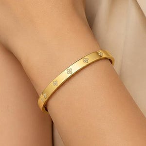 May include: A gold-toned metal bangle bracelet featuring a repeating pattern of small, four-petaled flower motifs. The surface of the bracelet is smooth and polished, with each flower design containing a tiny, clear, sparkling stone at its center.