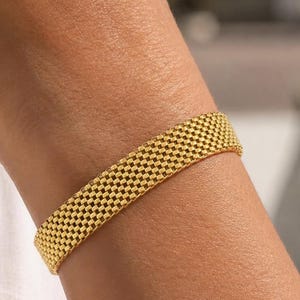 May include: A gold-toned bracelet featuring a dense, woven mesh texture with a repeating square pattern. The band has a uniform width and a flexible, linked construction that wraps around the wrist, showcasing a metallic finish and a consistent grid design.
