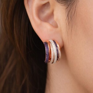 以下が含まれることがあります： 3連のローズゴールドフープピアス。1つのバンドは紫、1つは白、3つ目はローズゴールドです。ピアスには小さな輝く石がちりばめられています。耳に装着されています。