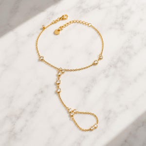 Könnte beinhalten: Ein goldfarbenes Handkettchen mit einem zarten Kettendesign, das mit mehreren klaren, rund geschliffenen Steinen besetzt ist. Das Schmuckstück verfügt über eine Ringkomponente für den Finger und einen Verschluss mit Verlängerungskette.