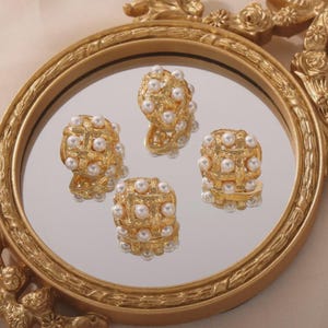 May include: Four gold-colored earrings with a woven design, each studded with small, white pearls. The earrings are displayed on a round, mirrored surface within a gold-framed decorative tray, creating a luxurious aesthetic.