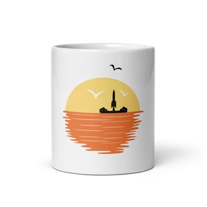 May include: White ceramic mug featuring a sunset design with a rocket launching into the sky. The design includes a yellow sun, orange water, and black silhouettes of a rocket and birds. A simple and modern design.