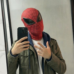 May include: A red and black Spider-Man mask with large black eye lenses. The mask is worn with a white shirt, blue hoodie, and olive green jacket. The person is holding a black smartphone.