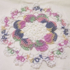 May include: A handmade, white crochet doily with a floral design. The doily features a central circular pattern surrounded by a ring of colorful, shell-shaped accents in pastel hues of pink, blue, yellow, and green. The outer edge has a scalloped trim.