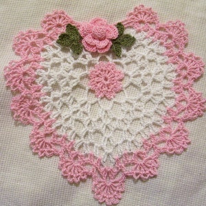 May include: A handmade, white and pink crocheted doily with a floral design. The doily features a central pink flower with green leaves and a smaller pink flower in the center. The outer edge is scalloped with pink crochet.