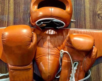 May include: A set of vintage boxing equipment in orange leather, including a head guard, gloves, and a protective cup. The gloves have white laces and trim. The equipment is displayed against a wooden background.