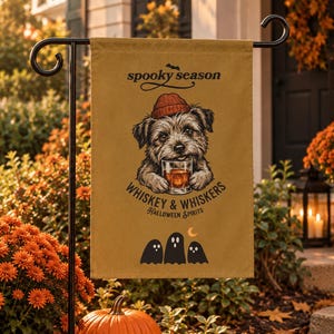 Op de afbeelding: Een beige tuin vlag met een afbeelding van een ruige hond met een roestkleurige muts die een glas met amberkleurige vloeistof vasthoudt. De tekst luidt spooky season, whiskey & whiskers en halloween spirits, met drie zwarte spooksilhouetten eronder.
