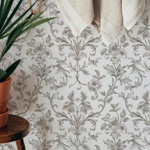 May include: A bathroom scene featuring a wooden towel rack with two beige towels, a woven net bag, and a natural bristle brush. The background is a floral wallpaper, and a potted plant sits on a wooden stool.