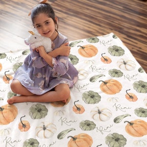 May include: A white blanket with a pattern of watercolor pumpkins in shades of orange, green, and white. The name "Remi" is written in cursive across the blanket. The blanket is on a wooden floor.