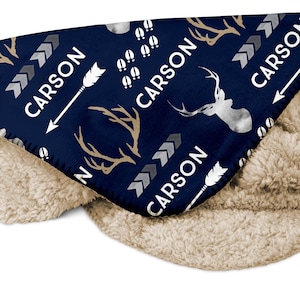 May include: A navy blue fleece blanket with a white and gold pattern of deer antlers, arrows, and footprints. The blanket has the name "CARSON" repeated throughout the pattern.