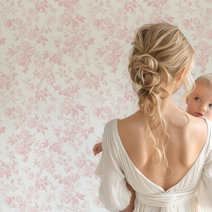 May include: A woman in a white dress holds a baby in her arms. The woman has long blonde hair and is wearing a white dress with a low back. The baby is wearing a white onesie. The background is a pink floral wallpaper.