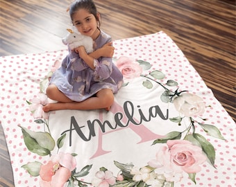 May include: A personalized blanket with the name "Amelia" in script lettering, surrounded by a floral wreath of pink roses and green leaves. The blanket has a white background with pink polka dots.