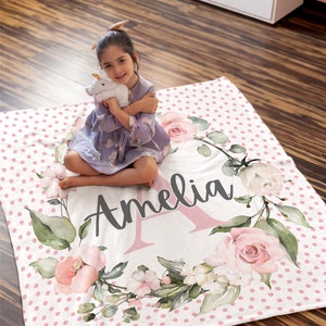 Puede incluir: Una manta personalizada con el nombre "Amelia" en escritura cursiva, rodeada por una corona floral de rosas rosas y hojas verdes. La manta tiene un fondo blanco con lunares rosas.