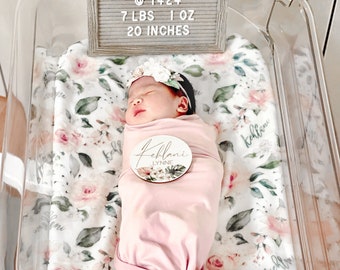 May include: A newborn baby girl swaddled in a pink blanket, lying in a clear plastic bassinet. The baby is wearing a white flower headband and a personalized wooden name tag that reads "Kehlani Lynne". A sign above the bassinet reads "Kehlani Lynne Peregrino August 11 2021 @ 1424 7 LBS 1 OZ 20 INCHES".