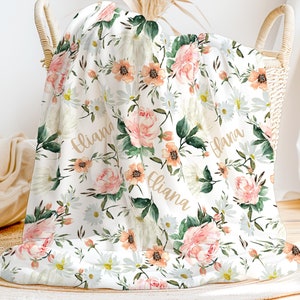 May include: A white floral baby blanket with a peach and green floral pattern. The blanket has the name "Eliana" written three times in a gold script font.