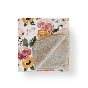 May include: A floral blanket with a watercolor design featuring sunflowers and peonies in shades of pink, orange, and yellow. The name "Lucy" is written in cursive. The blanket has a soft, beige sherpa lining.