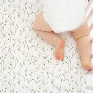 May include: A white crib sheet featuring a repeating pattern of delicate light pink flowers with thin green stems and leaves. The fabric is stretched over a mattress surface, showing a soft floral botanical print on a clean white background.