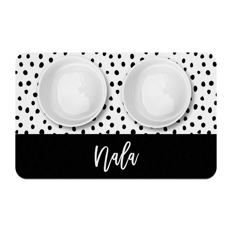 May include: Black and white polka dot pet food mat with two white ceramic bowls and the name "Nala" printed in white on a black strip.