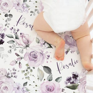 May include: A white crib sheet featuring a repeating pattern of light purple roses, small dark purple flowers, and green foliage. The name Rosalie is printed in a dark purple cursive script throughout the floral design on the fabric.