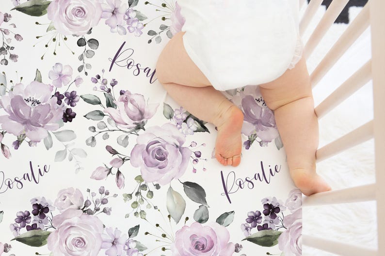 May include: A white crib sheet featuring a repeating pattern of light purple roses, small dark purple flowers, and green foliage. The name Rosalie is printed in a dark purple cursive script throughout the floral design on the fabric.