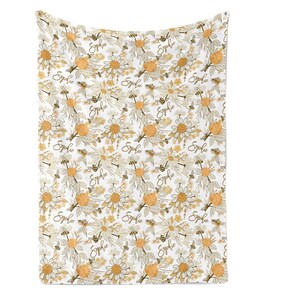 May include: A white blanket featuring a repeating pattern of yellow and white daisies, brown bees, and the name Sophie written in a brown cursive script. The design includes botanical elements with leaves and stems in a soft, muted color palette.