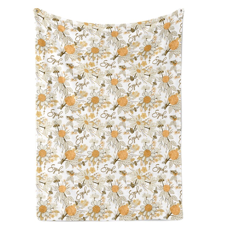 May include: A white blanket featuring a repeating pattern of yellow and white daisies, brown bees, and the name Sophie written in a brown cursive script. The design includes botanical elements with leaves and stems in a soft, muted color palette.