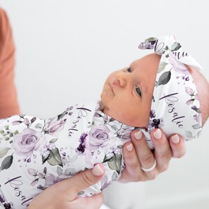 May include: A white swaddle blanket and matching headband featuring a floral pattern with purple roses, green leaves, and the name Rosalie printed in a cursive script. The fabric has a soft texture and is wrapped around a newborn infant.