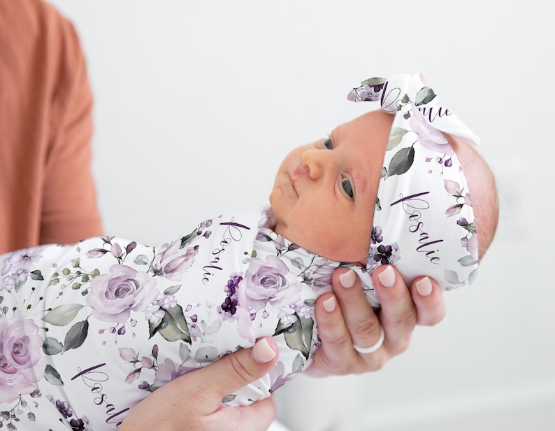 May include: A white swaddle blanket and matching headband featuring a floral pattern with purple roses, green leaves, and the name Rosalie printed in a cursive script. The fabric has a soft texture and is wrapped around a newborn infant.