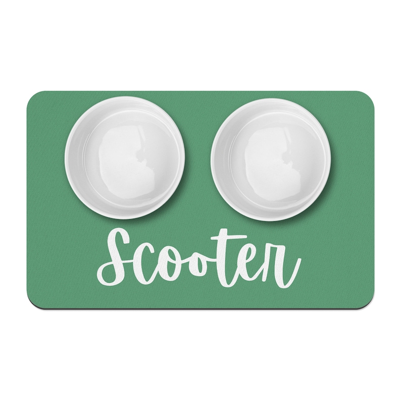 Puede incluir: Alfombra verde para comida de mascotas con dos cuencos de cer&aacute;mica blanca y el nombre "Scooter" escrito en cursiva blanca.