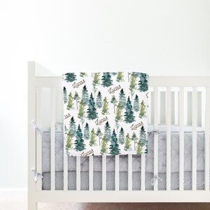 May include: A white crib with a gray and white striped crib skirt. A white baby blanket with a watercolor forest print and the name "Lucas" repeated throughout the blanket is draped over the crib.