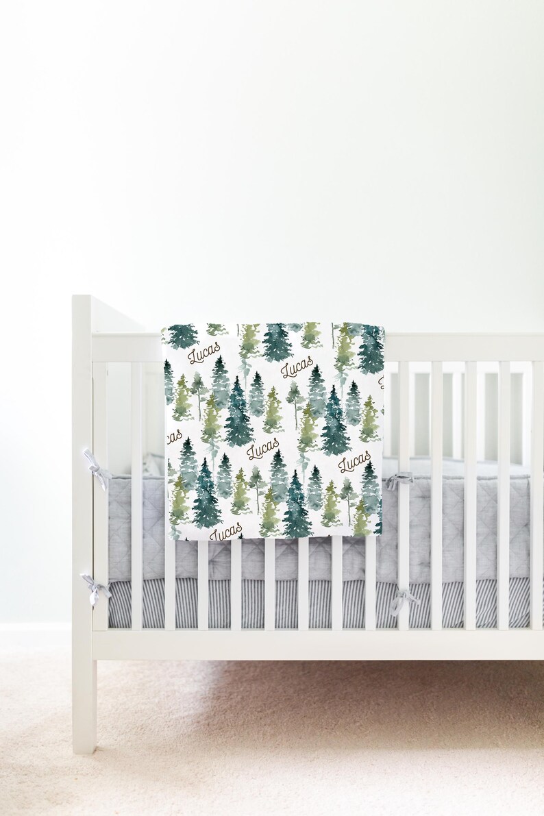 May include: A white crib with a gray and white striped crib skirt. A white baby blanket with a watercolor forest print and the name "Lucas" repeated throughout the blanket is draped over the crib.