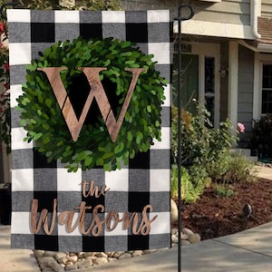 May include: A black and white buffalo check garden flag with a green wreath and a wooden letter W in the center. The text "the Watsons" is printed below the wreath.