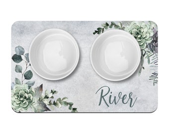 Pode incluir: Um tapete para tigelas de animais de estimação cinza e branco com um design floral verde e o nome "River" impresso nele. O tapete tem duas tigelas de cerâmica branca.