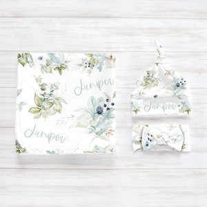 May include: A baby gift set with a white swaddle blanket, a white hat, and a white headband. The blanket and hat feature a watercolor floral pattern with green leaves and blue flowers. The headband has a bow and a floral pattern. The name "Juniper" is written on the blanket, hat, and headband.