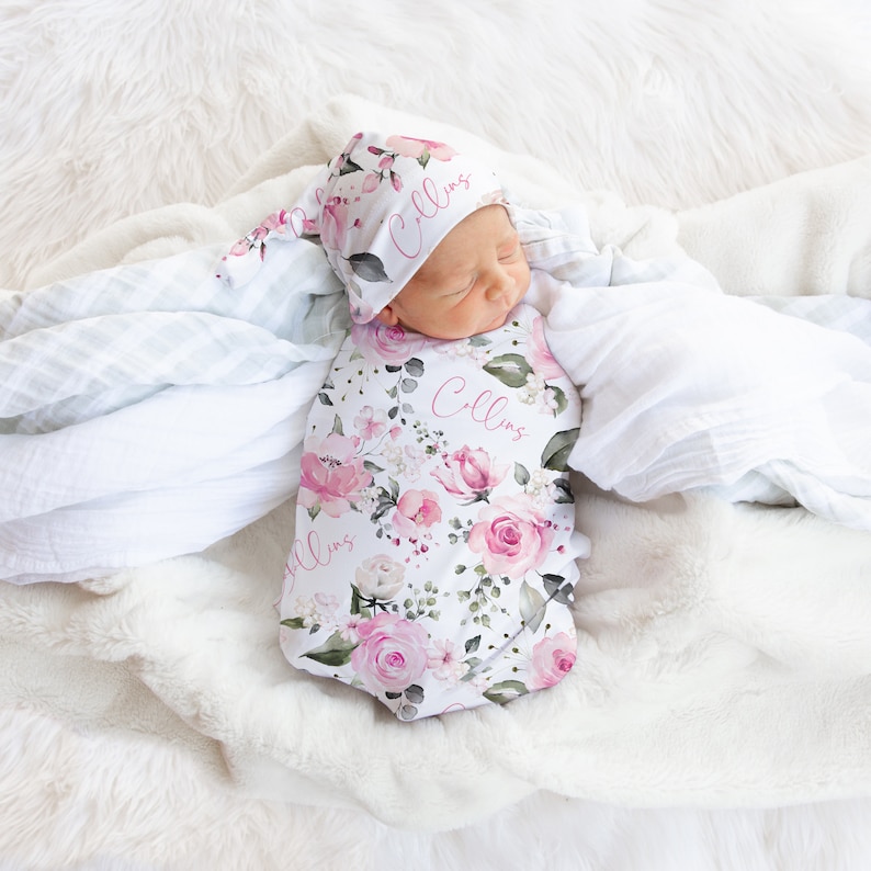 May include: A baby swaddle and matching hat set featuring a floral pattern with pink roses and green foliage. The name "Collins" is printed on both items in a pink script. The set is on a white, fluffy blanket.