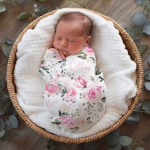 May include: A newborn baby swaddled in a floral print blanket, nestled in a woven basket. The blanket features pink roses and the name "Collins" in pink script. The basket is lined with a white, textured fabric.