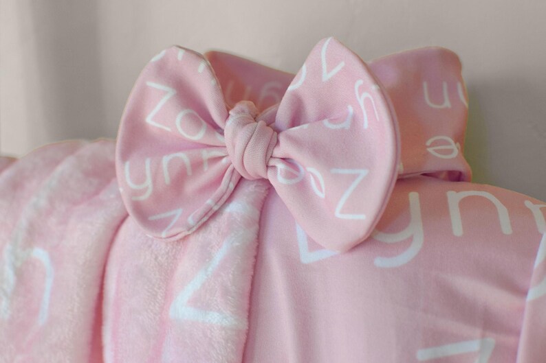 May include: A pink baby blanket with a white bow and white letters printed on it. The letters are the alphabet.