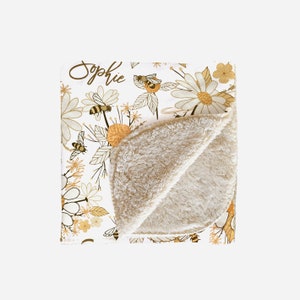 May include: A white throw blanket featuring a repeating pattern of yellow and brown bees, daisies, and wildflowers. The name Sophie is printed in brown script in the upper left corner. The blanket has a folded corner revealing a tan sherpa lining.